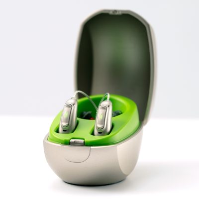 A pair of hearing aids stands upright and ready for use in an open Phonak Mini Charger Case with a green finish against a plain, light background.